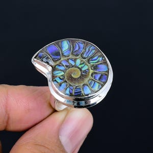 Könnte beinhalten: Ein Silberring mit einem polierten Ammonitenfossil. Das Fossil zeigt ein Spiralmuster mit schillernden Blau-, Grün- und Lilatönen. Der Ring hat ein schlichtes, poliertes Silberband und eine Fassung, die die natürliche Schönheit des Edelsteins hervorhebt.