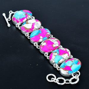 Könnte beinhalten: Ein silbernes Armband mit ovalen Steinen in Rosa, Blau, Weiß und Gold. Die Steine sind in Silber gefasst und durch eine Kette verbunden. Das Armband hat einen Knebelverschluss. Schmuck.
