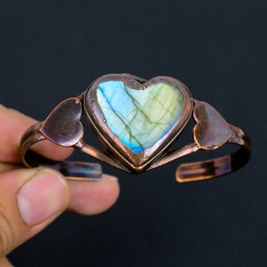 May include: A copper bracelet with a heart-shaped labradorite gemstone in the center. The bracelet has two smaller heart-shaped copper accents on either side of the gemstone.