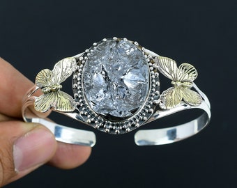 May include: Silver cuff bracelet featuring an oval, textured, clear stone center. The bracelet has gold-colored butterfly accents on each side of the stone. The bracelet is held against a dark background.