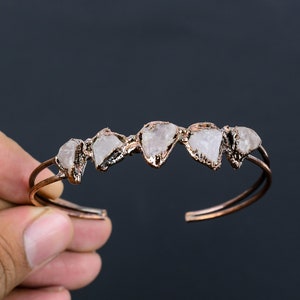 May include: A copper-colored cuff bracelet featuring five raw, light-colored gemstones. The gemstones are set in a copper-colored metal, creating a unique and natural look. The bracelet is open-ended, allowing for easy wear.