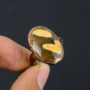 Op de afbeelding: Ovale ring met een grote, gepolijste steen in bruin- en geeltinten. De steen is gezet in een koperkleurige metalen band met een getextureerde rand. De ring wordt gepresenteerd tegen een donkere achtergrond.