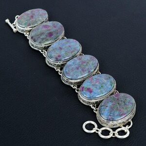 May include: A bracelet featuring six oval-shaped cabochons of ruby in zoisite, each set in a decorative silver filigree bezel. The stones display a mottled pattern of deep magenta, blue-grey, and green. The piece includes a toggle clasp closure.
