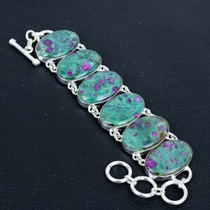Könnte beinhalten: Silberarmband mit sechs ovalen, grünen Edelsteinen mit rosa Einschlüssen. Die Steine sind in Silberfassungen gefasst und durch Silberketten verbunden. Das Armband hat einen Knebelverschluss.