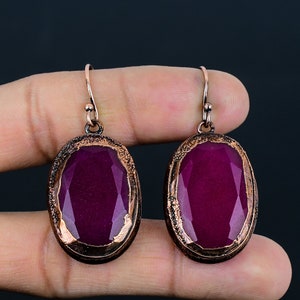 Peut inclure: Une paire de boucles d'oreilles ovales avec une pierre précieuse violette foncée sertie dans un cadre en métal ton cuivre. Les boucles d'oreilles ont une fermeture simple à crochet.