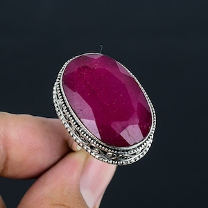 May include: A silver ring featuring a large, oval-shaped ruby gemstone. The ruby is a deep red colour and has a faceted cut. The ring has a decorative silver band with intricate details.