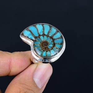 May include: A silver ring featuring a polished ammonite fossil. The fossil has a striking spiral pattern in shades of turquoise and brown, set in a silver band. The ring is a unique piece of jewelry.