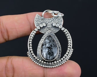 May include: A silver pendant with a cat-like design. The pendant features a teardrop-shaped stone with a black and white pattern, surrounded by a beaded circle and a textured top. The pendant is held by a finger.