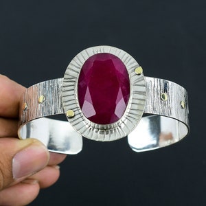 May include: A silver cuff bracelet featuring a large, oval-shaped ruby gemstone set in a silver and gold bezel. The bracelet has a textured surface and gold accents.