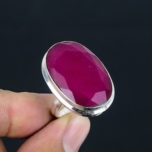 Op de afbeelding: Een zilveren ring met een grote, ovaal geslepen robijn. De robijn heeft een rijke, karmozijnrode kleur. De ring heeft een eenvoudige zilveren band, die de edelsteen benadrukt. De ring wordt tegen een donkere achtergrond gehouden.