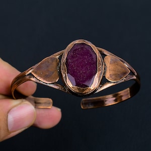 Può includere: Un braccialetto in rame con una grande pietra preziosa ovale di colore viola scuro incastonata al centro. Il braccialetto ha due tagli a forma di cuore su entrambi i lati della pietra preziosa.