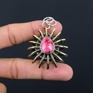 May include: A pendant with a teardrop-shaped, pink and white stone at the center, surrounded by a sunburst design. The pendant has silver and gold-colored metal accents and a silver loop for a chain.