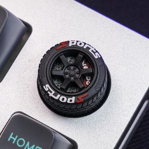 May include: A black and red sports car tire keycap with the word "Sports" printed on it. The keycap is sitting on a silver keyboard.
