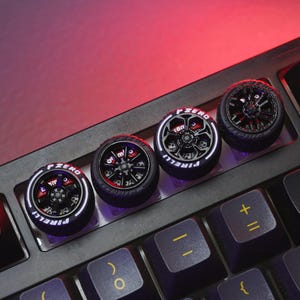 Puede incluir: Cuatro teclas negras de teclado con forma de neumáticos con la inscripción "P ZERO" y "PIRELLI" en blanco sobre un teclado negro. Las teclas de teclado están diseñadas para parecerse a neumáticos de coche.