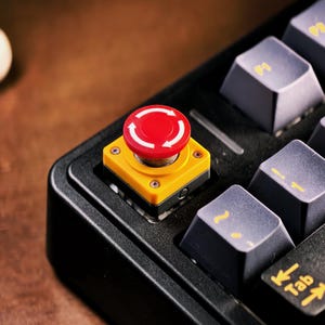 Peut inclure: Gros plan sur un clavier mécanique noir avec des touches grises et un bouton d'arrêt d'urgence rouge et jaune vif. Le bouton comporte des flèches blanches indiquant un mouvement circulaire. Le clavier est sur une surface en bois.