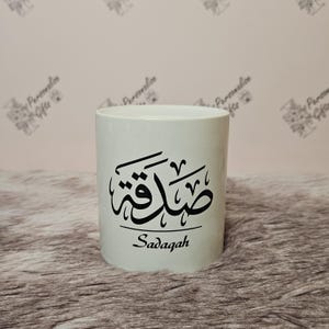 Peut inclure: Une tasse en céramique blanche avec une calligraphie arabe noire qui dit "Sadaqah" sur le devant.