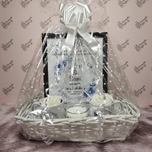 Puede incluir: Una cesta de mimbre blanca llena de un conjunto de regalo para una boda u ocasión especial. El conjunto incluye una impresión enmarcada con diseños florales azules y los nombres "Jenna & Sami" y la fecha "04/01/21". La impresión también incluye texto árabe. La cesta también incluye cuatro tazas blancas con diseños florales azules, dos velas blancas y papel de seda blanco.