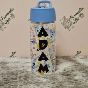 Peut inclure: Une bouteille d'eau en plastique transparent avec un couvercle bleu et une paille. La bouteille a un motif sur le th&egrave;me de l'espace avec des fus&eacute;es, des plan&egrave;tes et des &eacute;toiles. Le nom "ADAM" est imprim&eacute; en noir sur la bouteille.