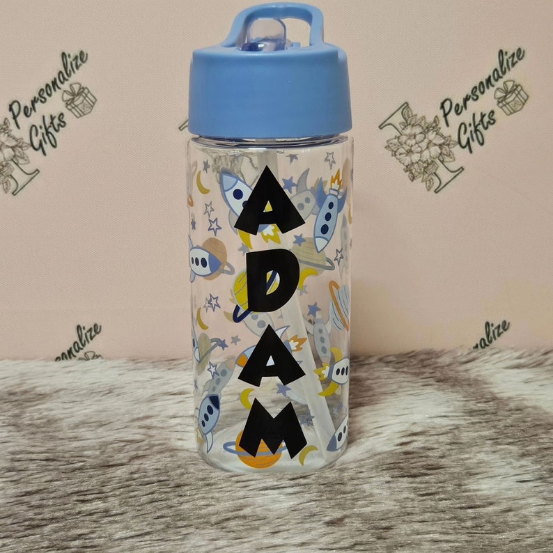 Peut inclure: Une bouteille d'eau en plastique transparent avec un couvercle bleu et une paille. La bouteille a un motif sur le th&egrave;me de l'espace avec des fus&eacute;es, des plan&egrave;tes et des &eacute;toiles. Le nom "ADAM" est imprim&eacute; en noir sur la bouteille.