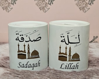 May include: Two white ceramic piggy banks with gold Arabic calligraphy and a gold silhouette of a mosque. The piggy bank on the left says "Sadaqah" and the piggy bank on the right says "Lillah".