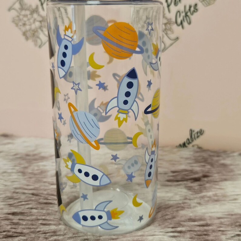 Peut inclure: Un gobelet en verre transparent avec une paille et un motif sur le th&egrave;me de l'espace. Le gobelet pr&eacute;sente des fus&eacute;es, des plan&egrave;tes, des &eacute;toiles et des lunes dans des tons de bleu, jaune et orange.
