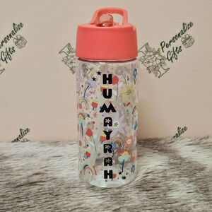 Peut inclure: Une bouteille d'eau en plastique transparent avec un couvercle rose et une sangle noire. La bouteille a un motif color&eacute; avec des arcs-en-ciel, des fleurs et des &eacute;toiles. Le nom "HUMAYRAH" est &eacute;crit verticalement sur la bouteille.