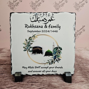 Könnte beinhalten: Eine weiße Schiefertafel mit goldener Umrandung und einem floralen Design. Die Tafel zeigt ein Bild der Kaaba und der Moschee in Mekka. Der Text auf der Tafel lautet "Umrah Mubarak", "Rukhsana & family", "September 2024/1446" und "Möge Allah SWT deine Umrah annehmen und all deine Gebete erhören".