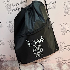 May include: A black drawstring backpack featuring a front zippered pocket. The bag is printed with white Arabic text, the year 2026, a graphic of the Kaaba, and the name Safa in a script font. It includes black cord straps and metal eyelets.
