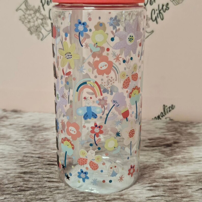 Peut inclure: Une bouteille d'eau en plastique transparent avec un couvercle rose et une paille. La bouteille est d&eacute;cor&eacute;e d'un motif color&eacute; de papillons, de fleurs et d'arcs-en-ciel.