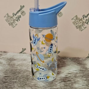 Peut inclure: Une bouteille d'eau en plastique bleu et transparente avec un couvercle &agrave; rabat et une paille. La bouteille a un design sur le th&egrave;me de l'espace avec des fus&eacute;es, des plan&egrave;tes et des &eacute;toiles.