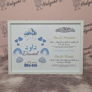 Op de afbeelding: Een ingelijste print met een witte rand toont blauwe en witte illustraties van wolken, regenbogen en een trein. De naam "Dawood" is zowel in het Arabisch als in het Engels geschreven. Er staat ook tekst in Arabisch schrift.