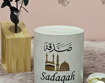 May include: White ceramic piggy bank with a gold mosque design and the Arabic word "Sadakah" written in black. The text "Sadakah" is written in English below the mosque design.