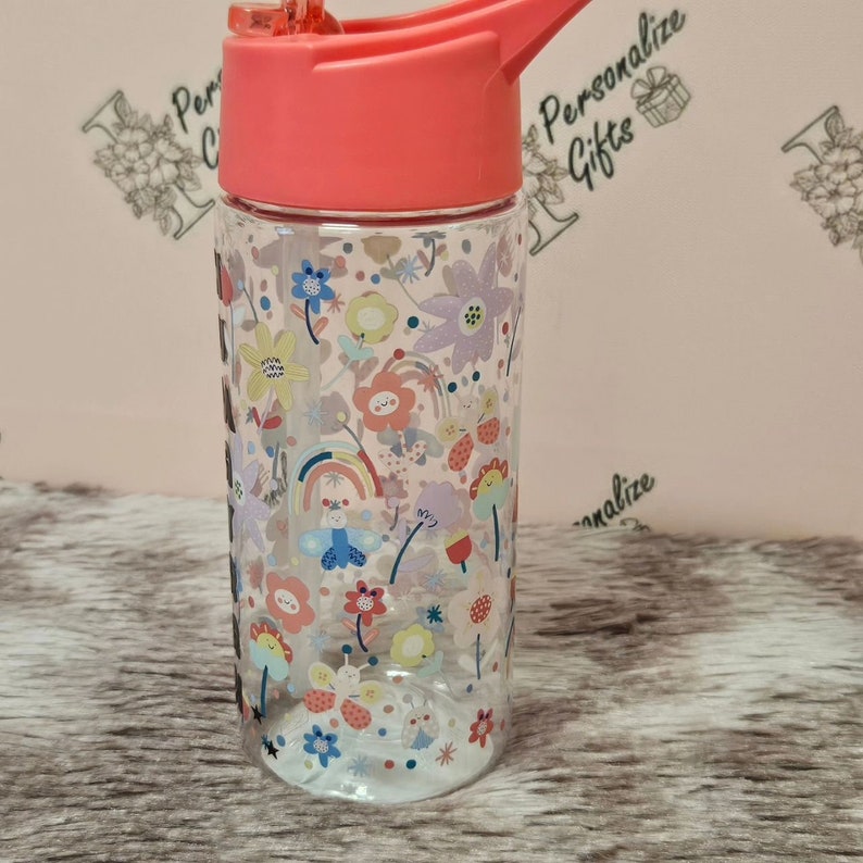Peut inclure: Une bouteille d'eau en plastique transparente avec un couvercle rose et une paille. La bouteille est d&eacute;cor&eacute;e d'un motif color&eacute; de fleurs, d'arcs-en-ciel et de papillons. Le texte "Personalize Gifts" est visible en arri&egrave;re-plan.