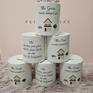 Peut inclure: Cinq tirelires en céramique blanche avec un motif de maison et le texte "The Gora Family Sadaqah Jar" et "The Soni Family Sadaqah Jar" dessus. Les tirelires ont une citation sur la charité dessus.