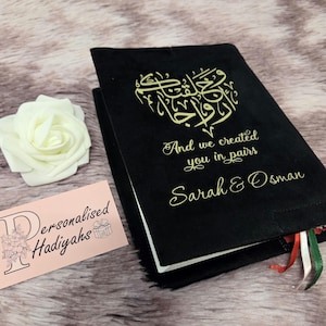 May include: Black book with gold calligraphy in a heart shape, with the text "And we created you in pairs" and the names "Sarah & Osman". A white rose and a pink business card are next to the book.
