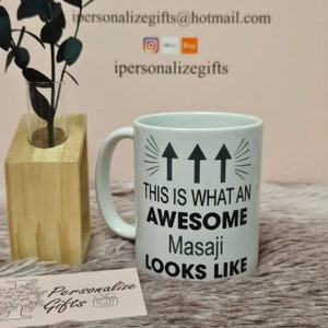 以下が含まれることがあります： 黒いデザインで「This is what an awesome Masaji looks like」と書かれた白いセラミック製のコーヒーカップ。上向きに3本の黒い矢印があります。