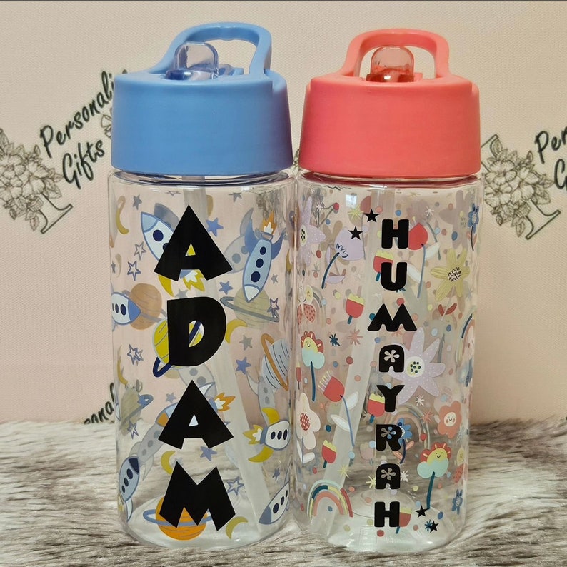 Peut inclure: Deux gourdes personnalis&eacute;es avec des pailles et des couvercles. La gourde de gauche a un couvercle bleu et un motif sur le th&egrave;me de l'espace avec le nom "ADAM" imprim&eacute; dessus. La gourde de droite a un couvercle rose et un motif floral avec le nom "HUMAYRAH" imprim&eacute; dessus.