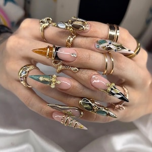 Könnte beinhalten: Eine Hand, die lange, mandelförmige Nägel mit aufwendigen Designs und Goldschmuck zeigt. Die Nägel weisen eine Vielzahl von Verzierungen auf, darunter goldene Akzente, Edelsteine und detaillierte Nagelkunst. Goldringe schmücken die Finger und ergänzen die aufwendigen Nageldesigns.