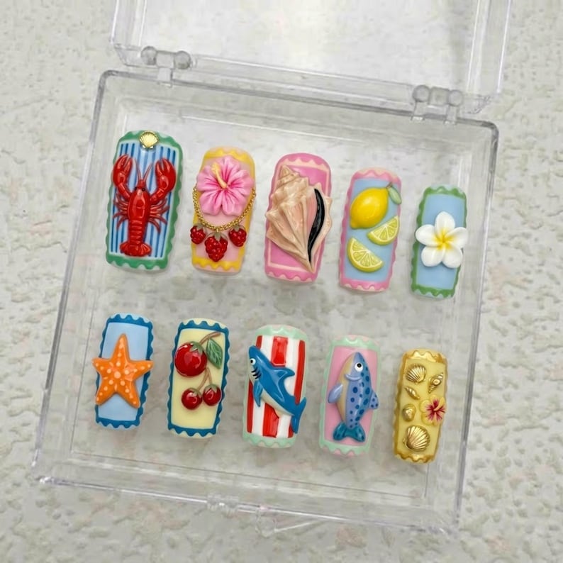 May include: A set of ten press-on nails with a summer theme. Each nail features a different design, including a red lobster, pink flower, seashell, lemon slices, starfish, cherries, shark, fish, and other tropical elements. The nails are in a clear plastic case.
