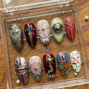 May include: A collection of ten artificial nails with diverse designs. The nails showcase metallic, jewel, and painted patterns, including a shield, sword, starburst, and geometric motifs. The nails are presented in a clear plastic case.