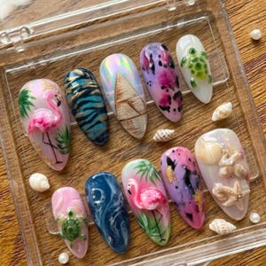Könnte beinhalten: Eine Sammlung von zehn künstlichen Fingernägeln mit verschiedenen Designs. Die Nägel zeigen tropische Themen, darunter Flamingos, Schildkröten, Muscheln und abstrakte Muster in Rosa, Blau und Lila. Die Nägel werden in einer transparenten Kunststoffbox präsentiert.