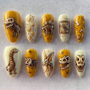 May include: A set of ten press-on nails with a dinosaur theme. The nails feature various designs, including dinosaur skeletons, footprints, and a dinosaur skull, in shades of yellow, brown, and white. The nails are oval-shaped and have a glossy finish.