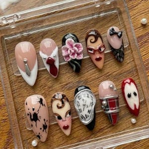 Könnte beinhalten: Eine Sammlung von zehn künstlichen Nägeln mit verschiedenen Designs. Die Nägel weisen eine Reihe von Farben auf, darunter Nude, Rosa, Schwarz und Rot, und sind mit aufwendigen Details wie floralen Akzenten, Gesichtern und abstrakten Mustern verziert. Die Nägel werden in einer transparenten Kunststoffbox präsentiert.