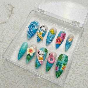 May include: A set of ten long, almond-shaped artificial nails with a tropical theme. Designs include blue waves, Snoopy surfing, flowers, and a pink flamingo. The nails are displayed in a clear plastic case.