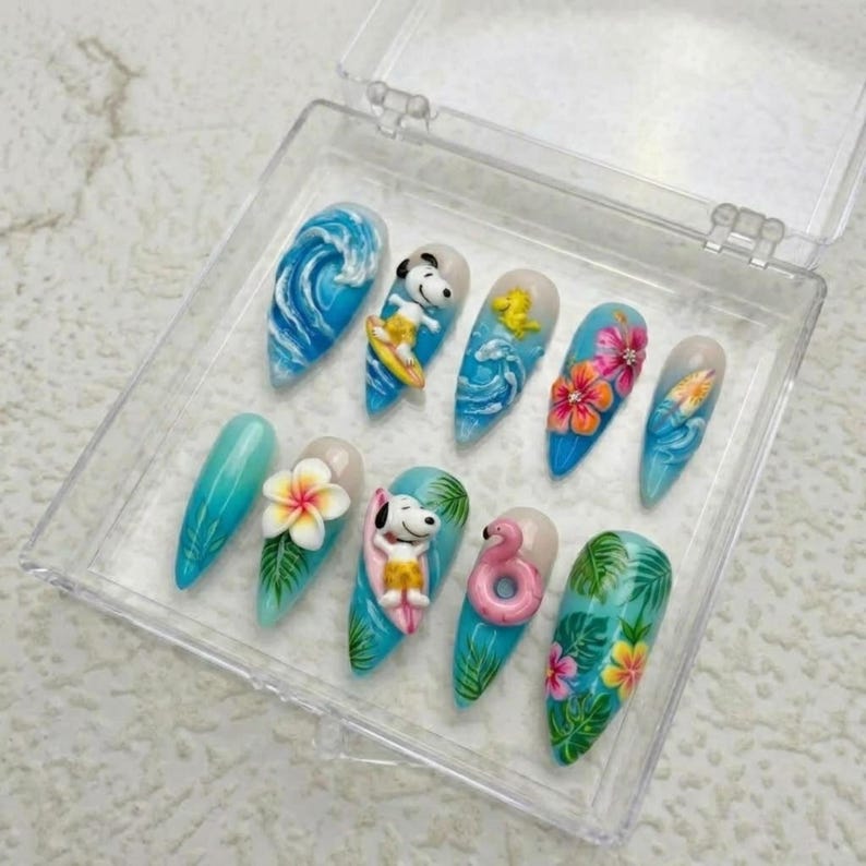 May include: A set of ten long, almond-shaped artificial nails with a tropical theme. Designs include blue waves, Snoopy surfing, flowers, and a pink flamingo. The nails are displayed in a clear plastic case.