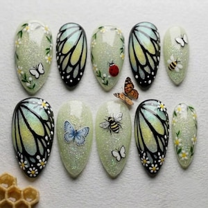 May include: A set of ten almond-shaped press-on nails featuring a light green glitter base. Designs include black butterfly wing patterns, small white daisies, a red ladybug, a bumblebee, and individual blue, white, and orange butterflies.