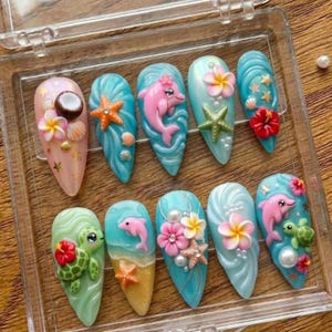Könnte beinhalten: Ein Set aus zehn langen, mandelförmigen Kunstnägeln mit tropischem Thema. Jeder Nagel hat ein anderes Design, darunter Delfine, Seesterne, Muscheln und Blumen, in den Farben Rosa, Blau und Grün. Die Nägel sind in einer transparenten Kunststoffbox angeordnet.