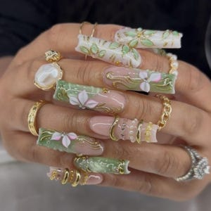 May include: A hand displaying long, ornate acrylic nails. The nails feature a combination of pink, white, and green designs with gold accents. Some nails have 3D floral embellishments and gold rings.