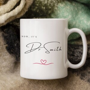 Könnte beinhalten: Weiße Keramiktasse mit einer schwarzen handschriftlichen Aufschrift "Now, it's Dr. Smith" mit einem roten Herzen und einer roten Linie darunter.