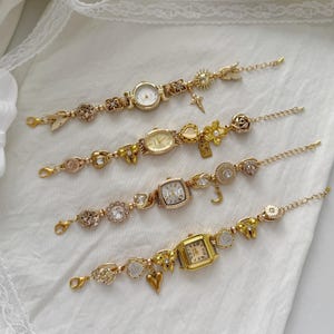 May include: Four gold-toned charm bracelets with various watch faces and decorative charms. The bracelets feature round, oval, and square watch faces, adorned with hearts, moons, and floral accents. The bracelets are displayed on a white surface.
